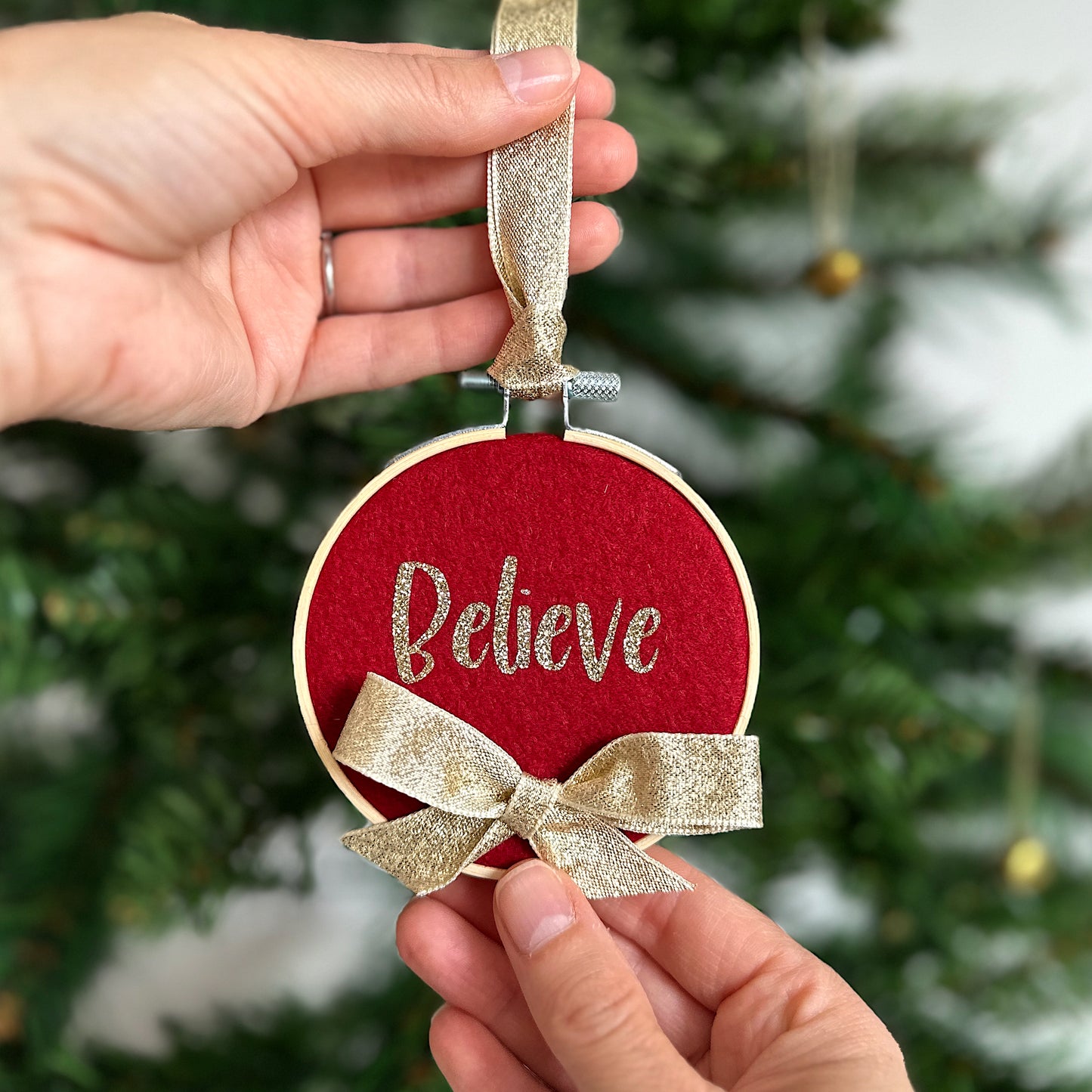 Red ornament with gold bow and 'Believe' text held by hands against a Christmas tree background