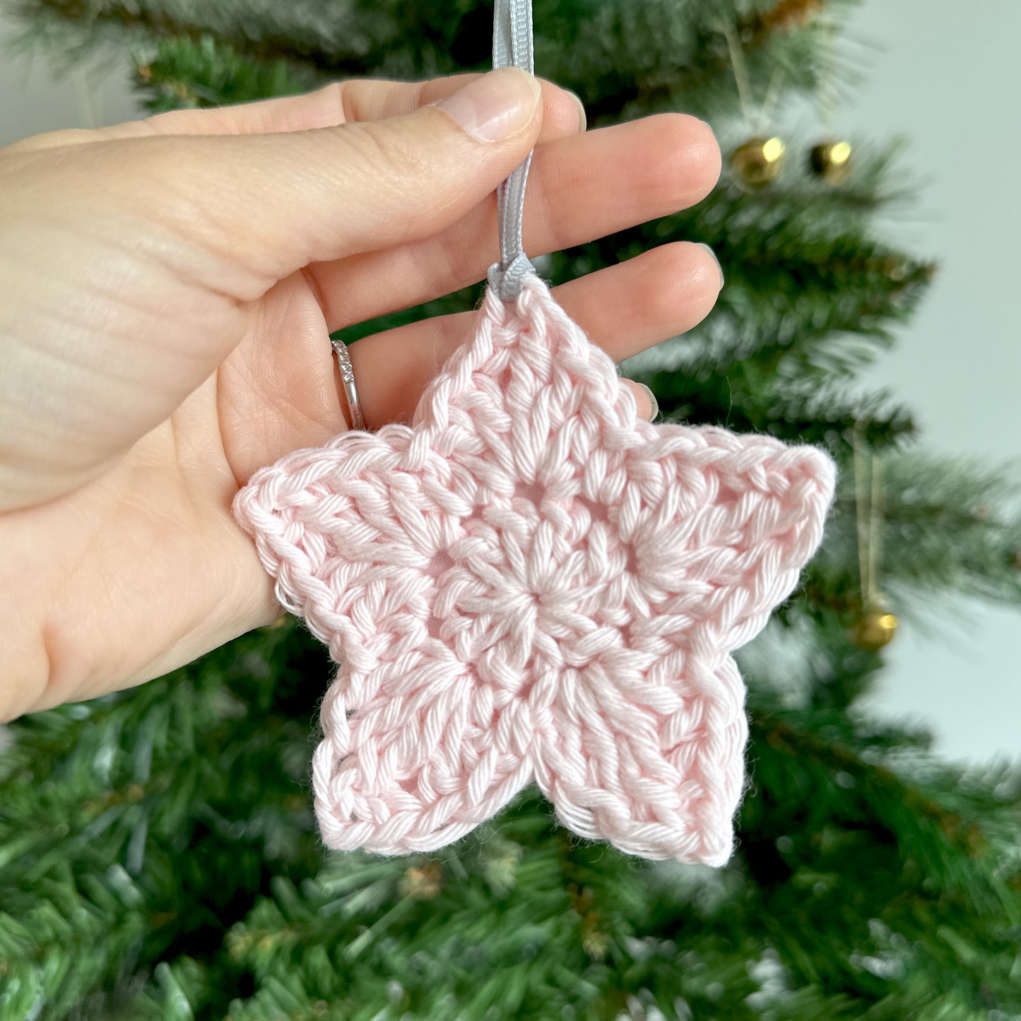 Pink crocheted star ornament held in front of a Christmas tree.