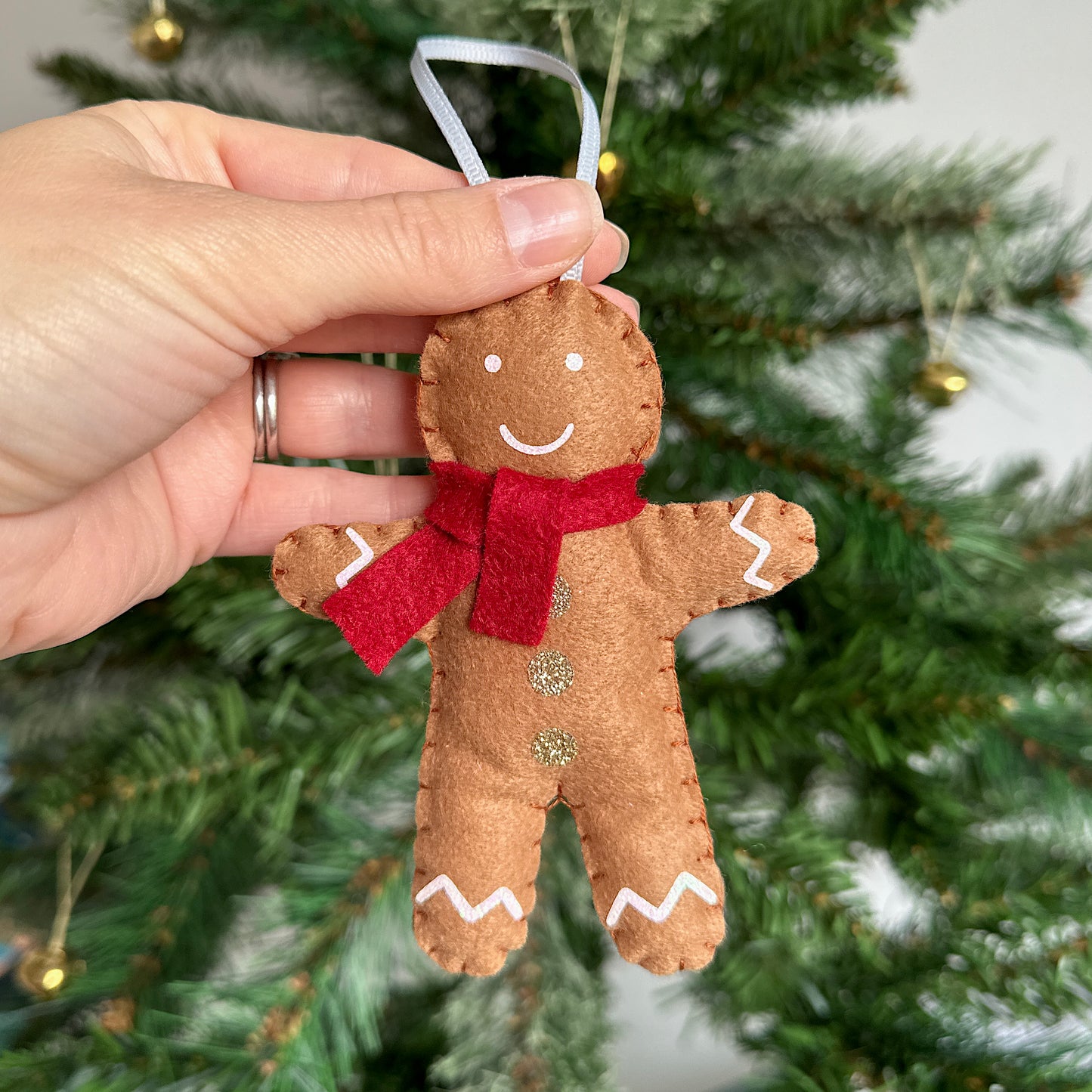 Hand holding a gingerbread man decoration with a red scarf in front of a Christmas tree