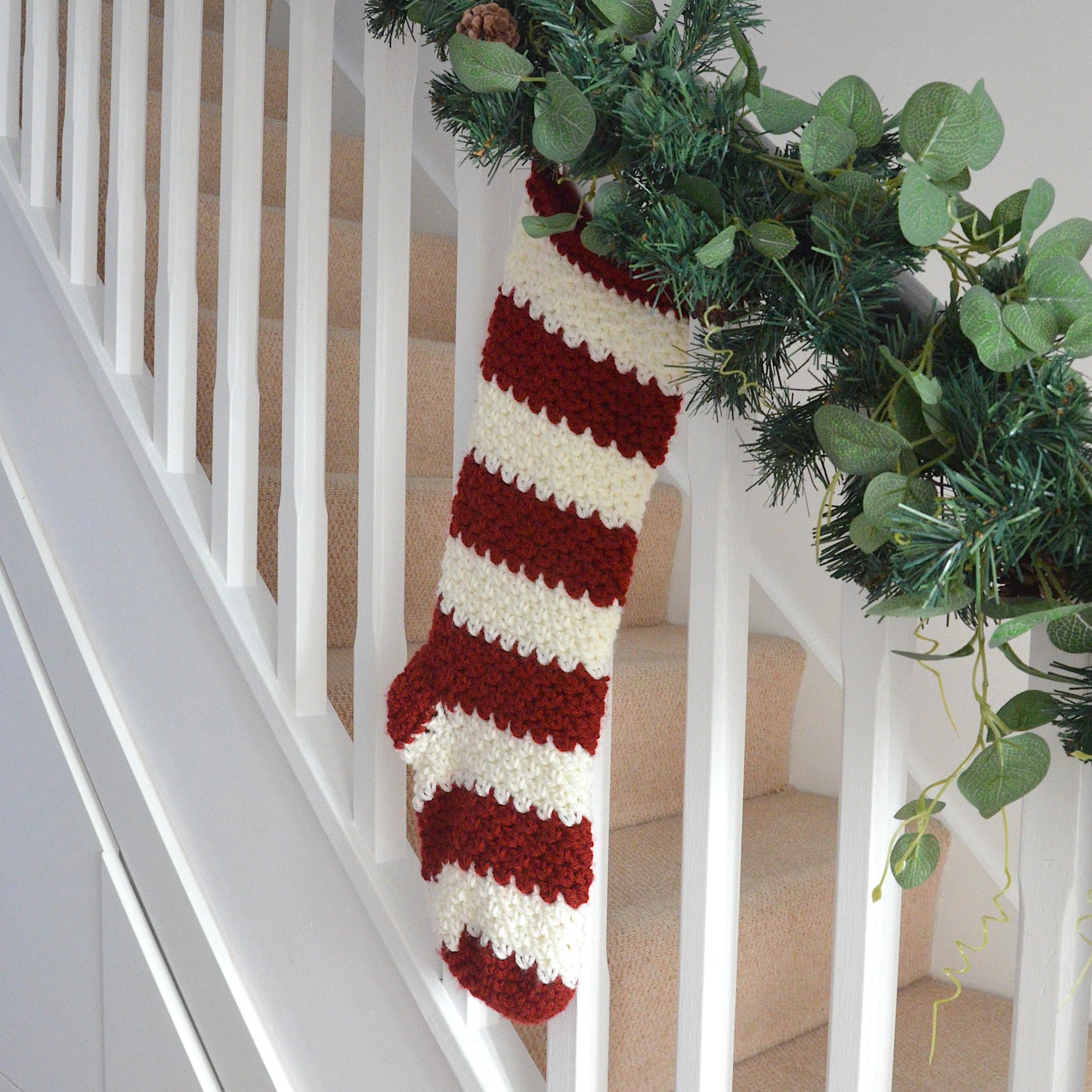 Red and white striped Christmas stocking hanging on a staircase with garland decoration.