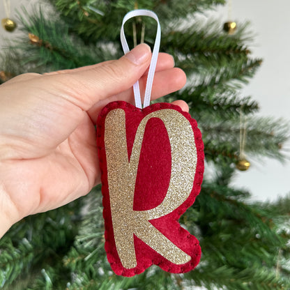 Red and gold letter ornament held in front of a Christmas tree.