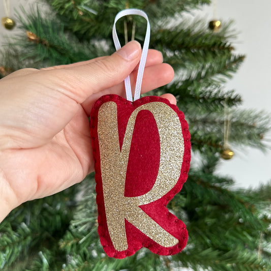 Red and gold letter ornament held in front of a Christmas tree.