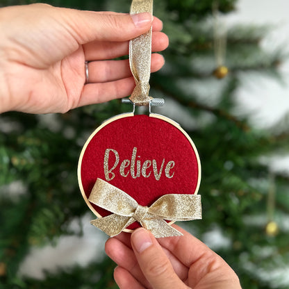 Red ornament with gold bow and 'Believe' text held by hands against a Christmas tree background